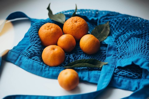 Fresh tangerines with green leaves arranged in a vibrant blue string bag, set against a white table