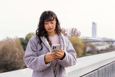 Young Latin woman in a casual jacket checking her phone outdoors, standing on a bridge with trees
