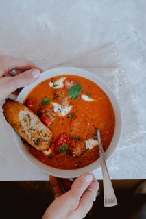 From above cropped unrecognizable person holding a bowl of tomato soup with meatballs, bread, and