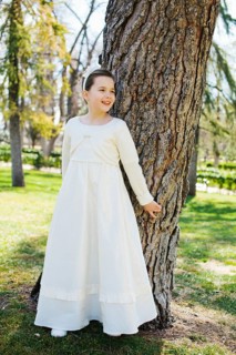 A young girl stands by a tree, smiling in a beautiful white First Communion dress. Sunlight filters