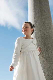 A young girl wearing a white First Communion dress stands outdoors against a stone column. The
