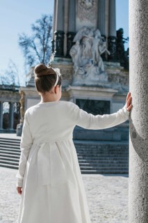 A young girl wearing a white First Communion dress stands beside a stone column, with a historic