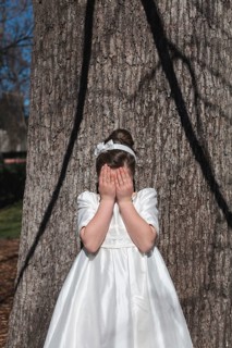 A young girl stands in front of a tree, covering her face with her hands. She is wearing an elegant