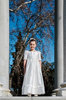 A young girl stands in a white First Communion dress, framed by stone columns and bare trees, with