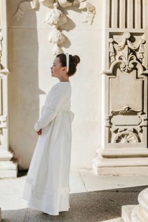 A young girl wearing a First Communion dress stands by an ornate wall. The scene captures a serene