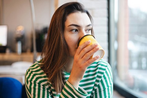 A young woman in a striped shirt takes a peaceful coffee break, sipping from a yellow cup inside a