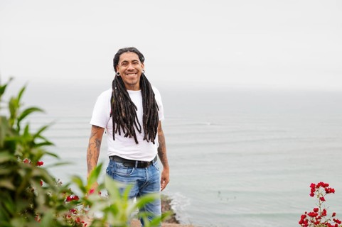 A man with long dreadlocks and tattoos smiles warmly, standing near vibrant flowers with the serene