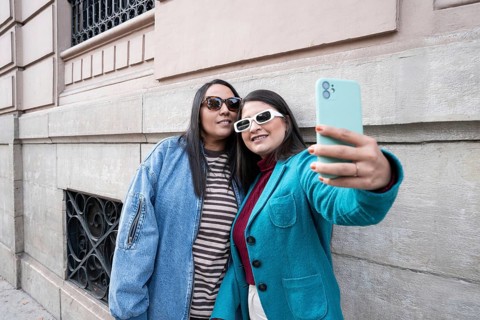 A Latin lesbian couple takes a joyful selfie outdoors, leaning against a historic building. Both