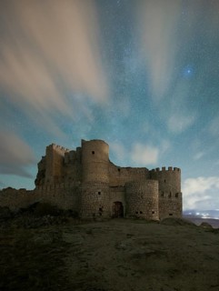 The medieval Manqueospese Castle in Avila, Spain, stands under a starry sky, with ethereal clouds