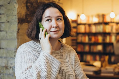 A young woman in a cream sweater smiles while talking on her green smartphone inside a warm, cozy