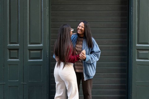 A joyful Latin lesbian couple stands in an embrace, laughing together in front of a green door.
