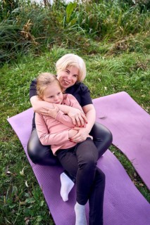 A joyful mother and daughter sit together on a yoga mat outdoors in a riverside. The lush, green