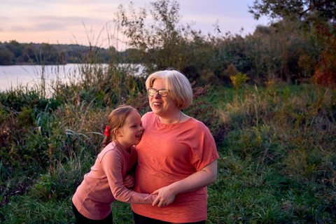 A joyful mother and daughter share a laugh in a serene outdoor setting by the riverside at sunset.
