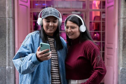 A latin lesbian couple listens to music with headphones while looking at a smartphone outdoors.
