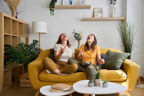 A joyful lesbian couple sits on a vibrant yellow sofa, tossing popcorn in the air and laughing