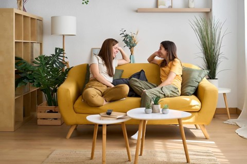 A cheerful lesbian couple enjoys a cozy moment on a yellow sofa The room is filled with greenery