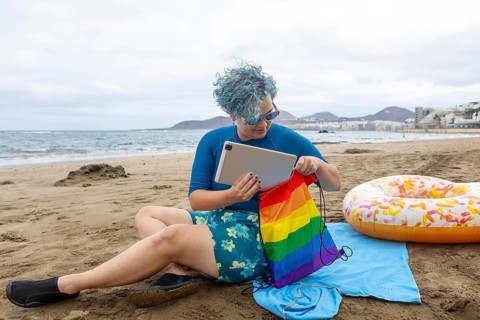 A transgender man with blue hair sitting on a beach, putting a tablet inside a rainbow pride bag.
