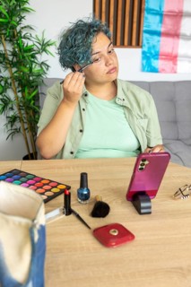 A transgender man meticulously applies makeup while recording a tutorial with his mobile phone.