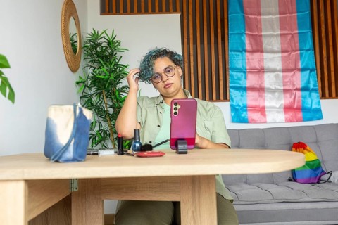 A transgender man sits at a wooden table, engaging in a makeup tutorial using his mobile phone.