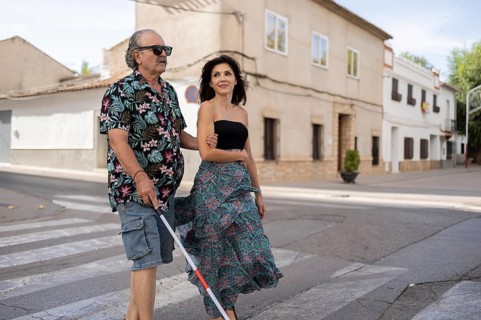 A blind father uses a white cane while his daughter guides him through a tranquil town street. Both