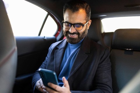 A happy Indian man, looking down at his smartphone, sits in a car, dressed in a winter coat and