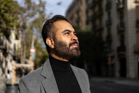 A confident Indian man dressed in a business-appropriate gray coat and black turtleneck is captured