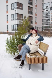 A couple sits on a wooden bench surrounded by snow, holding a small Christmas fir They are bundled