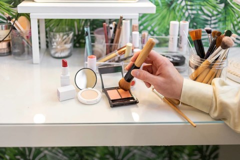 A hand uses a brush to apply organic makeup on a white vanity Various cosmetics, including lipstick