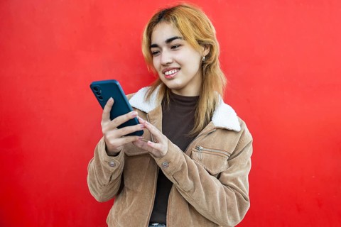 A Mixed-race woman smiles as she uses a smartphone, against a vibrant red background She is wearing