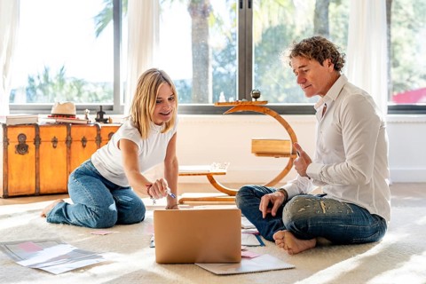 A man and woman sitting on the floor at home, engaging in work and planning activities. The man and