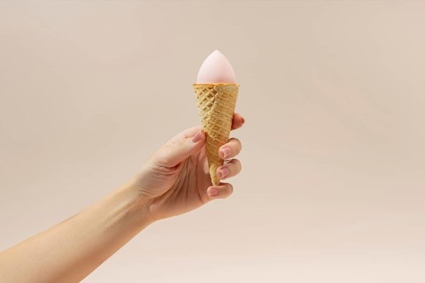 A hand holds a waffle cone with a pink cosmetic sponge inside, resembling ice cream The neutral
