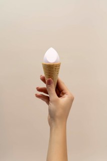 A hand holding a waffle cone with a cosmetic sponge inside, set against a soft, neutral background
