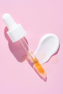 A dropper dispenses golden serum beside a white cream smear on a vibrant pink surface, representing