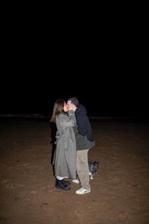 A couple shares a tender kiss under the night sky on a sandy beach, accompanied by a small dog The