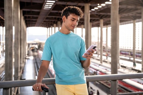 A young man in a blue t-shirt stands at a train station, holding a suitcase and using his