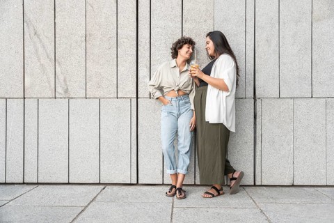 A relaxed lesbian couple enjoys a casual moment together, leaning against a modern stone wall. Both
