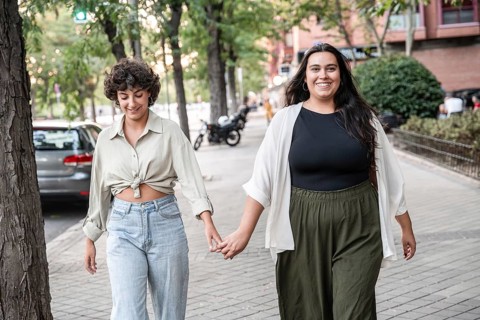 A lesbian couple is walking hand in hand on a city street, smiling and enjoying a sunny day. The