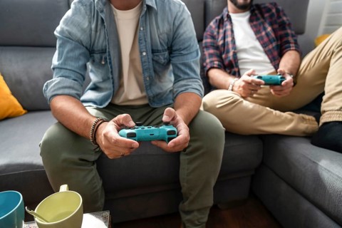 A gay couple sits on a couch, immersed in a video game. They both hold controllers, wearing casual