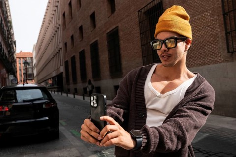 A stylish latin man wearing glasses and a yellow beanie takes a picture with his smartphone on a