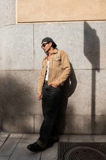 A man stands casually in front of a stone wall, wearing sunglasses, a flat cap, a beige jacket, and