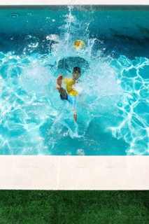 A young boy in a yellow shirt and blue shorts jumps energetically into a bright blue swimming pool,