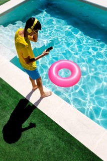 A person in vibrant attire enjoys music with headphones at a pool. Next to them, a pool float adds
