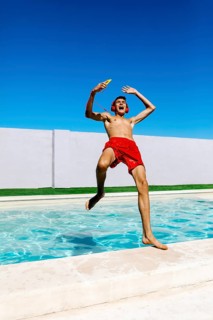 A joyful young teenage boy in red trunks leaps into a pool, wearing headphones and holding a