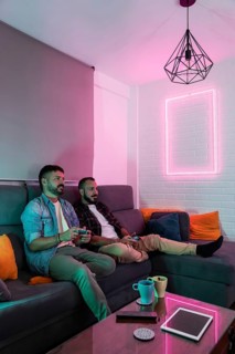 A gay couple relaxes on a sofa, playing video games in a warmly lit living space with modern decor.