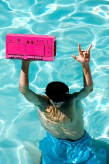 A person in blue shorts enjoys a sunny day in a pool, holding a vibrant pink boombox above the