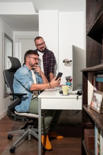 A gay couple collaborates at home, with one seated and the other standing, reviewing a smartphone.