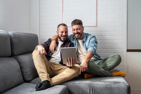 A joyful gay couple is comfortably seated on a sofa, engaged with a tablet. Their smiles reflect