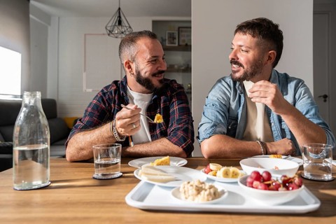 A happy gay couple sits together in a bright dining room, sharing a meal and enjoying each other's