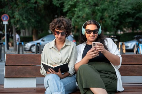 A lesbian couple enjoys a sunny day outdoors, sitting on a bench. One is reading a book, and the