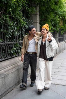 A joyful gay couple strolls arm in arm along a leafy city sidewalk, sharing smiles and enjoying